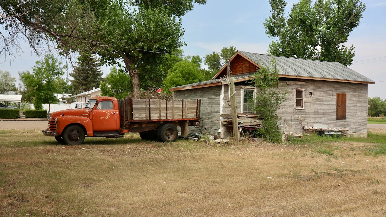 An abandoned house with a vintage orange truck parked beside it in a rural setting.